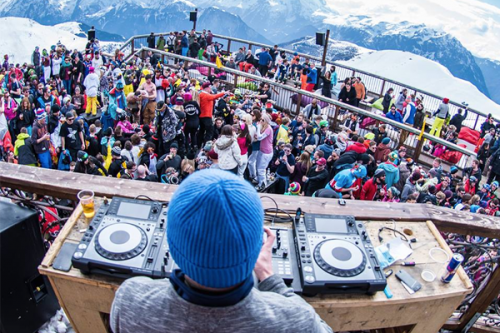 DJ with his deck in foreground looking out at crowd on a balcony with snowy mountains behind
